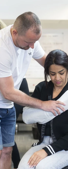 A man helps a seated woman by gently supporting her arm in a makeshift sling, practicing first aid skills together in a classroom setting. A man helps a seated woman by gently supporting her arm in a makeshift sling, practicing first aid skills together in a classroom setting.